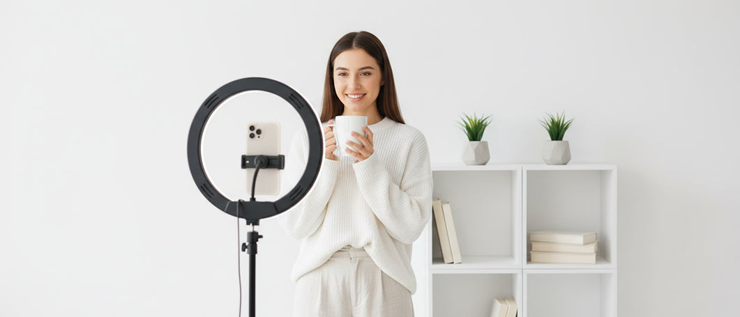 A woman in a white lounge set stands in a bright room holding a white mug, positioned in front of a black ring light with a smartphone mounted inside it. A white shelving unit with books and small potted plants is in the background.